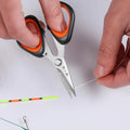 Hands using 110mm Zhang Xiaoquan AU fishing scissors to cut fishing line, featuring ergonomic grip and corrosion-resistant steel blades.