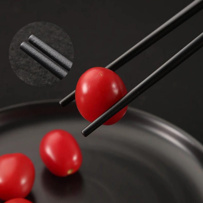 Non-slip black chopsticks holding a red cherry tomato on a black plate.