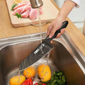 Chef's knife being washed under running water with vegetables in sink.