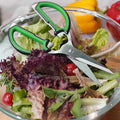 Kitchen scissors cutting fresh salad greens in a glass bowl.