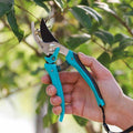 Hand holding 185mm pruning shears while trimming tree branches outdoors.